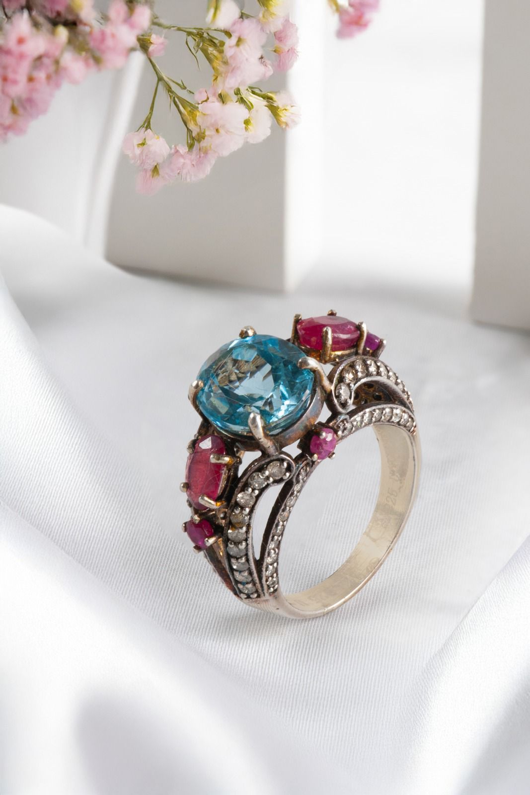 Close-up of Imperial Vintage Style Ring showcasing Blue Topaz center stone with deep red Thai Rubies and shimmering Melee Diamonds set in sterling silver, placed on white silk fabric.