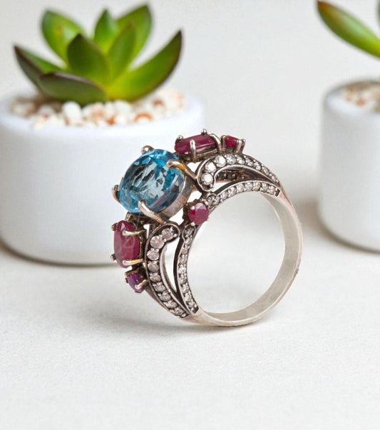 Handcrafted Imperial Vintage Style Blue Topaz Ring with Thai Rubies and Melee Diamonds in sterling silver, photographed elegantly beside green succulents.